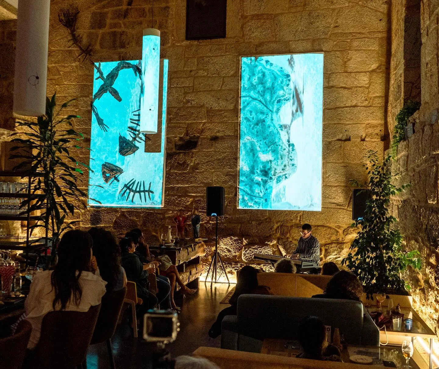 Concierto en el salón: público sentado en los sofás mientras un pianista toca bajo dos proyecciones de vídeo sobre la pared de granito