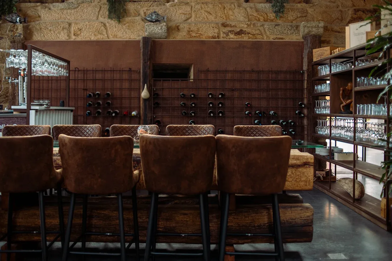 Pared del salón convertida en botellero: malla metálica oxidada con botellas y taburetes de cuero delante