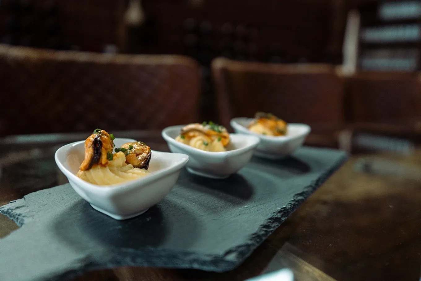 Tres cuencos blancos con mejillones sobre muselina de patata, servidos en una tabla de pizarra